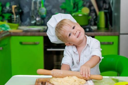 happy little boy preparing dough in kitchen at table. there are dough products on table, dressed as chef