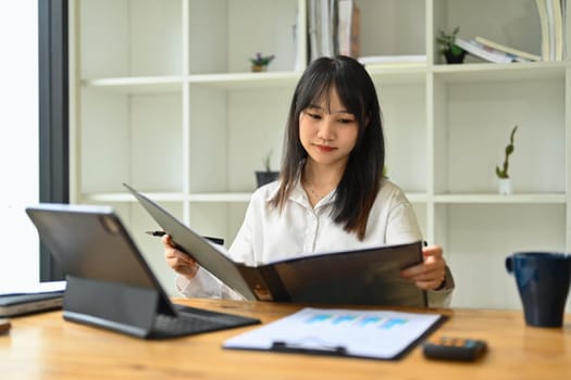 Concentrated asian female employee reviewing financial reports at her workplace.