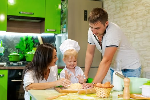 happy family mom and little son and dad are preparing dough in kitchen at table. products for dough are on table