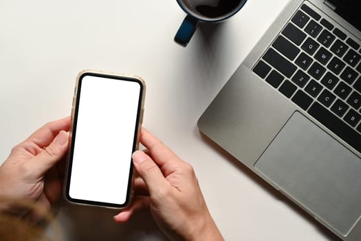 Overhead view of man holding smartphone over white office desk with laptop and coffee cup.