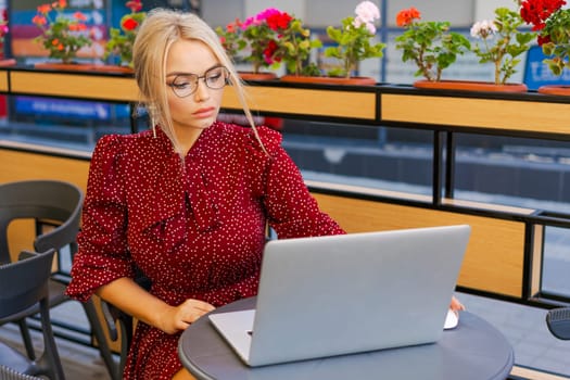 Beautiful woman works on laptop in coffee shop and using mobile phone in a red dress and glasses, business blonde online business