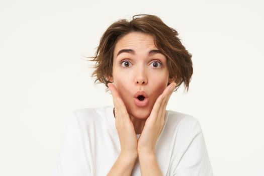 Portrait of girl with surprised face, makes shocked expression, stands over white background. Emotions and people concept