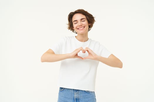 Portrait of young woman shows heart, love and care gesture, express sympathy, like something or someone, smiling happily, standing over white background.