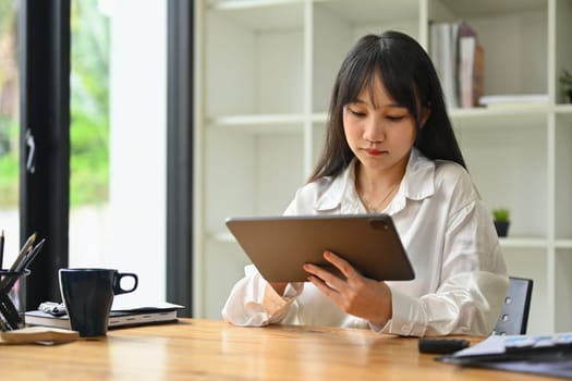 Image of pretty young female employee sitting in bright office and using digital tablet.