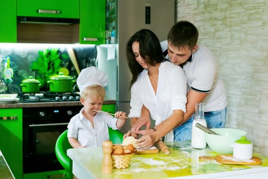 happy family mom and little son and dad are preparing dough in kitchen at table. products for dough are on table