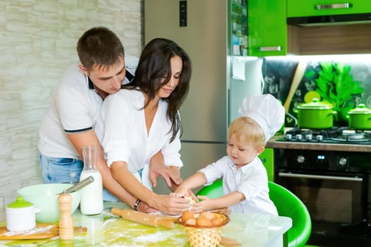 happy family mom and little son and dad are preparing dough in kitchen at table. products for dough are on table