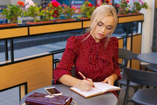 Nice caucasian woman writes in notebook spending time outdoors in cafe. Blonde girl wears a red dress. Inspiration concept and working with studies remotely