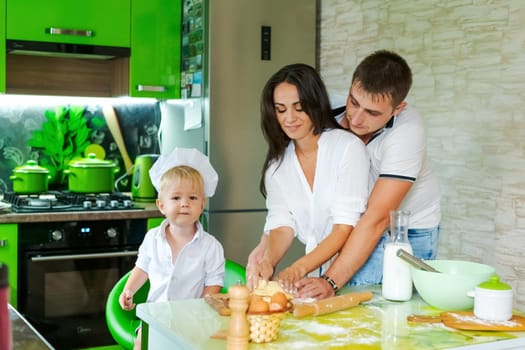 happy family mom and little son and dad are preparing dough in kitchen at table. products for dough are on table