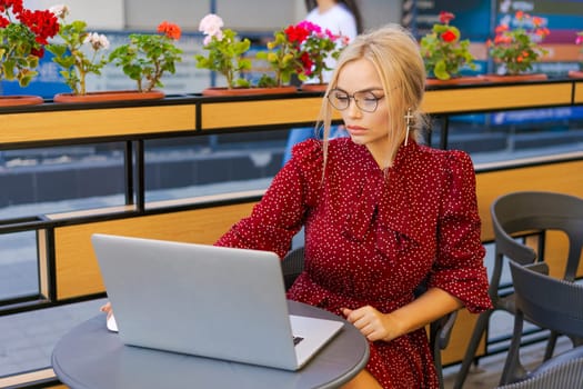 Beautiful woman works on laptop in coffee shop and using mobile phone in a red dress and glasses, business blonde online business