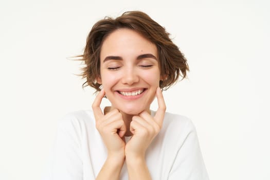 Portrait of brunette woman smiling, shuts her eyes and touches dimples on her cheeks, stands over white background. Copy space