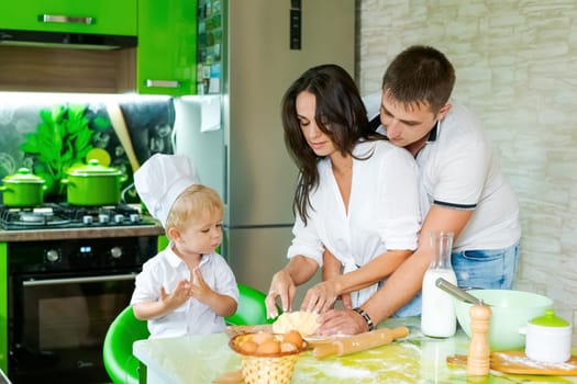 happy family mom and little son and dad are preparing dough in kitchen at table. products for dough are on table