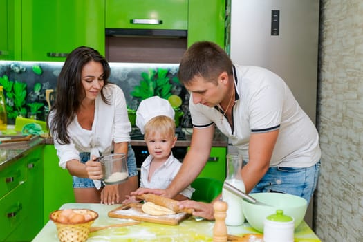 happy family mom and little son and dad are preparing dough in kitchen at table. products for dough are on table