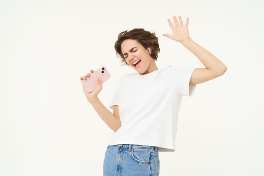 Portrait of carefree girl singing in wireless headphones into smartphone microphone, playing music game on mobile app, standing over white background.