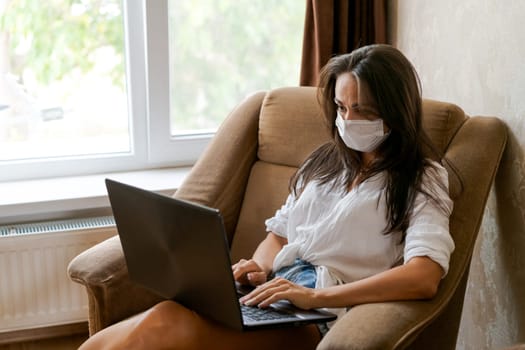 Cute woman at home in chair with laptop wearing protective mask on her face communicates on an online conference, concept of working from home, freelancer or study