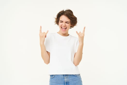 Happy and excited young woman shows horns gesture, heavy metal, having fun, posing over white background.