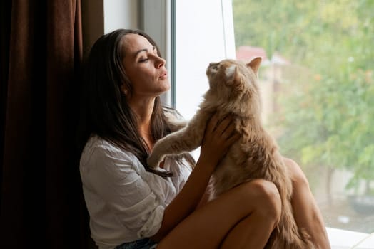 cute woman sitting on the windowsill with a red cat and looking out the window