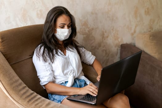 Cute woman at home in chair with laptop wearing protective mask on her face communicates on an online conference, concept of working from home, freelancer or study