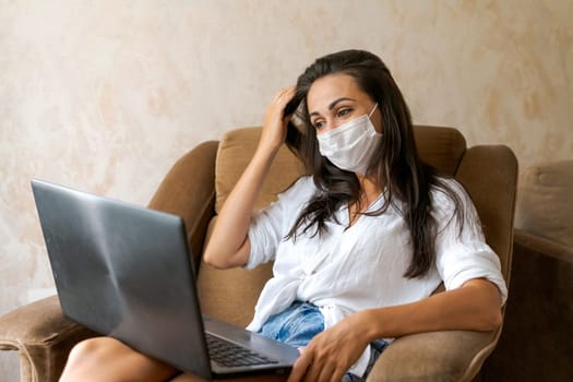 Cute woman at home in chair with laptop wearing protective mask on her face communicates on an online conference, concept of working from home, freelancer or study