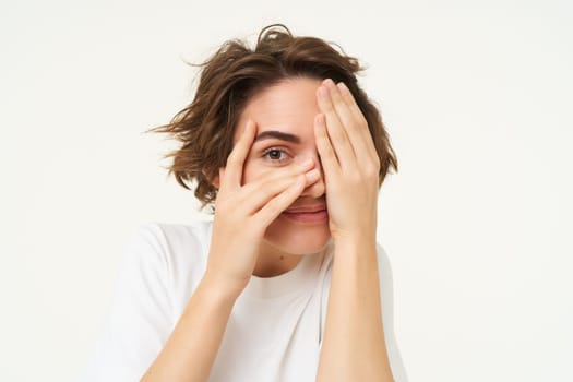 Happy brunette woman in white t-shirt, peeks through fingers, looks at surprise and smiling, stands against studio background.