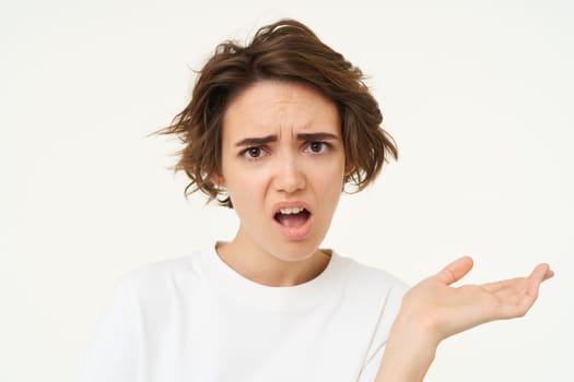 Portrait of arrogant young woman, raising one hand, looking disappointed, complaining, frowning and cringing, standing over white background. Copy space