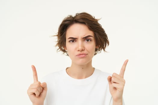 Sceptical young woman, pointing fingers up, frowning and sulking looking perplexed, unsure about something, standing over white studio background.