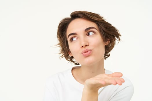 Close up of brunette woman blowing air kiss, mwah, kissing gesture, pucker lips and gazing away, standing over white background. Copy space