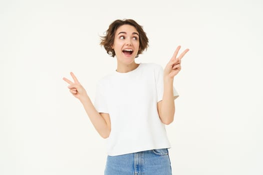 Portrait of young girl shows peace, v-sign and smiling, express posivity and joy, posing over white studio background.