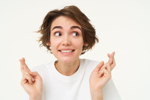 Close up of hopeful young woman, student hopes for dream come true, cross fingers for good luck, makes wish, stands over white background. Copy space