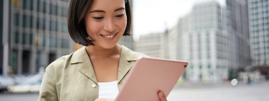Portrait of asian woman reading, using tablet while standing on street, smiling while looking at screen.