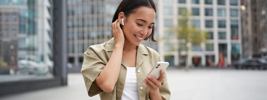 Smiling asian girl listens music in wireless headphones, looks at her phone, choosing music or podcast. Young woman calling someone, using headphones.