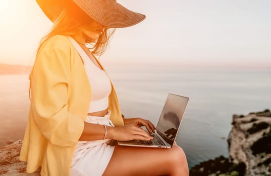 Successful business woman in yellow hat working on laptop by the sea. Pretty lady typing on computer at summer day outdoors. Freelance, travel and holidays concept.