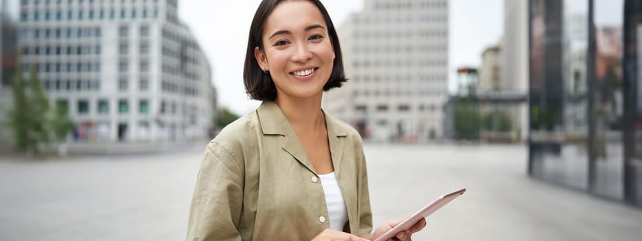 Young smiling asia woman with tablet, standing on street in daylight.