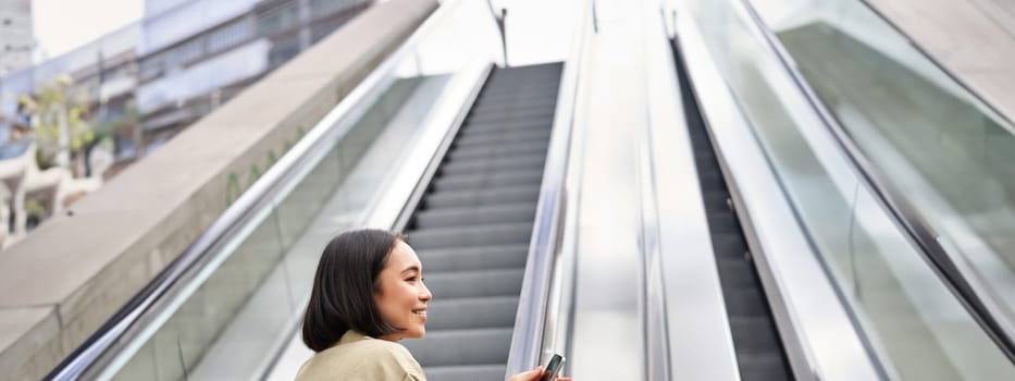 Young happy woman standing on escalator with smartphone, going up, walking in city.