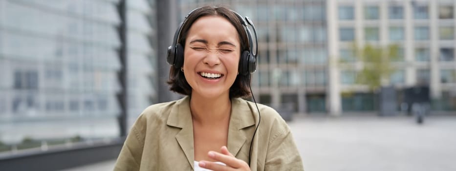 Carefree asian girl, laughing and smiling, wearing headphones and walking on street. Outdoor shot of young woman listening music and looking happy.