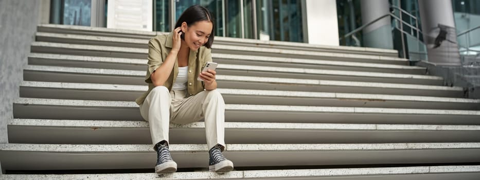 Cellular technology and people. Young happy asian girl sits with smartphone in front of building. Woman using mobile phone, smiling while looking at screen.