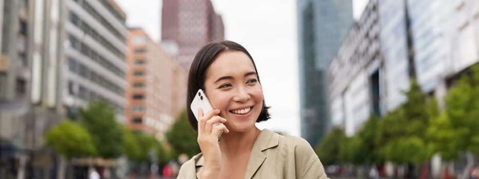 Portrait of smiling asian woman talking on mobile phone, walking on street near busy road and speaking to friend, laughing.