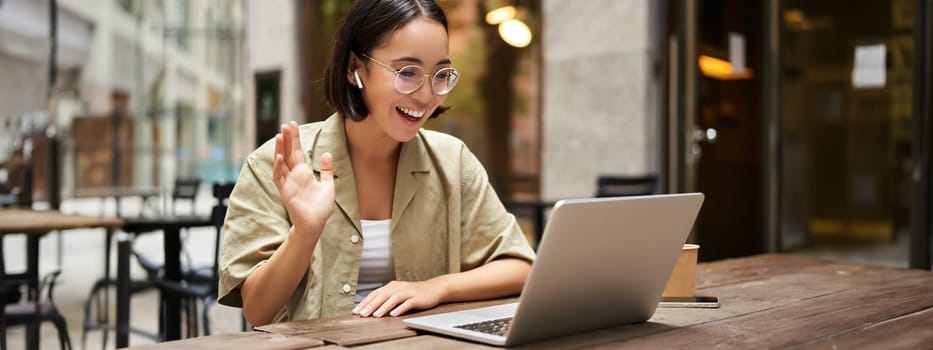 Portrait of young woman video chatting, having online meeting on laptop, sitting in outdoor cafe with compute, wearing glasses and smiling.