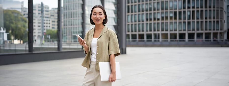 Asian girl with laptop and smartphone, standing on street of city centre, smiling at camera.