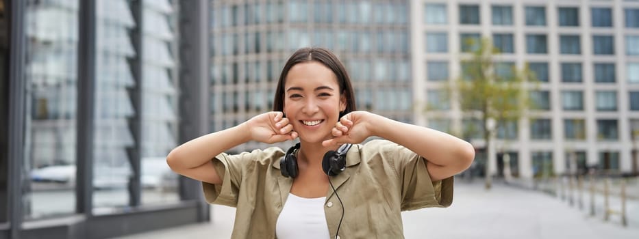 People. Portrait of smiling asian girl with headphones, dancing on street of city centre, looking happy.