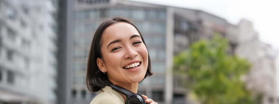 Vertical shot of beautiful asian woman posing with headphones around neck, smiling and laughing, standing on street in daylight.