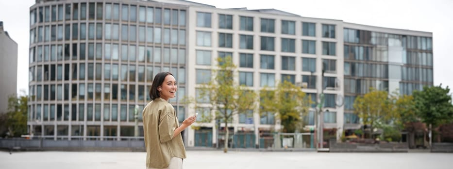 Silhouette of asian woman walking on street in wireless headphones, holding smartphone.