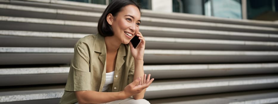 Portrait of beautiful asian girl talks on mobile phone, sits on street stairs. Woman with smartphone smiling, making a call.