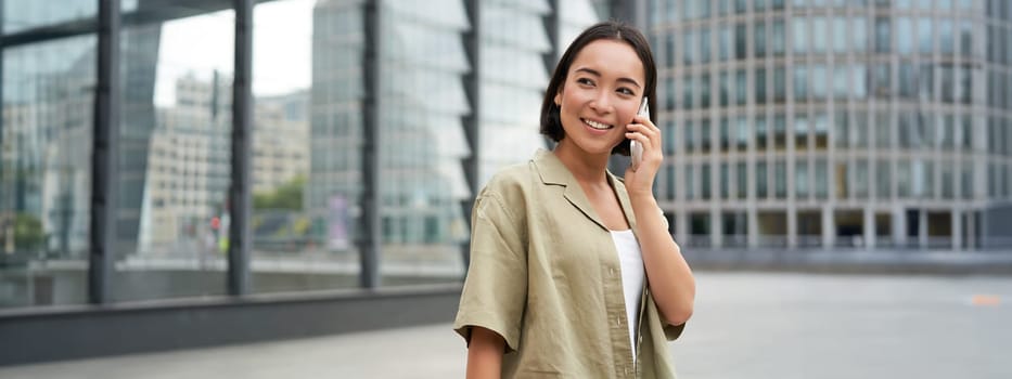 Portrait of asian girl smiling, talking on phone, making a call, standing on street near building and waiting for someone, answer telephone.