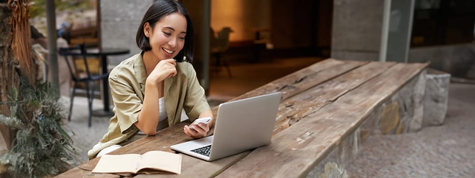 Portrait of stylish urban woman working on laptop from outdoor cafe, sitting with documents and computer, smiling.