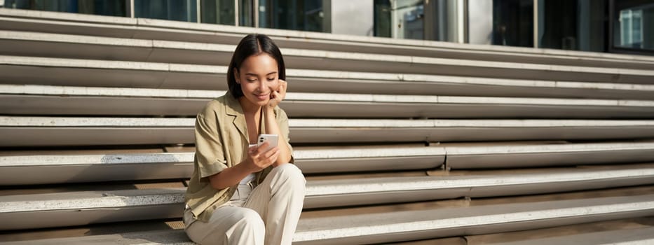 Vertical shot of asian woman, student sits on stairs in city, looking at mobile phone screen and smiling, using smartphone app.