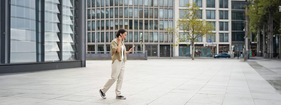 Silhouette of asian woman walking on street in wireless headphones, holding smartphone.