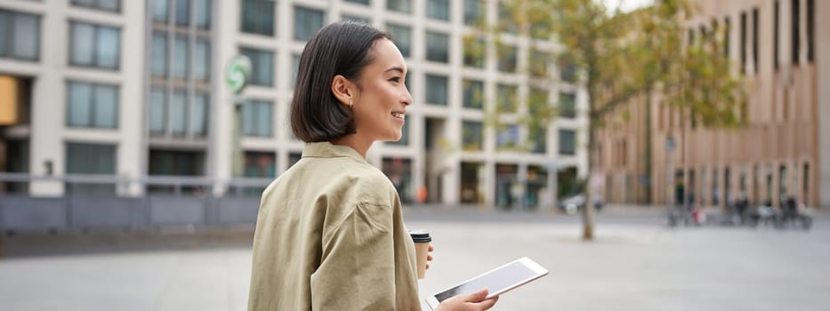 Asian girl walking on street, reading on tablet and drinking coffee outdoors.