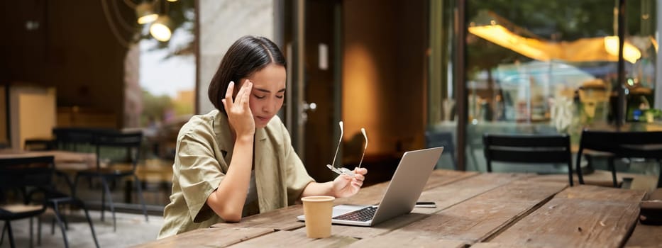 Young asian woman feeling tired after working with laptop, sitting in cafe on bench outdoors, drinking coffee, looking exhausted.