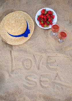 straw hat, wine and a plate of strawberries on a sandy beach-inscription love the sea.