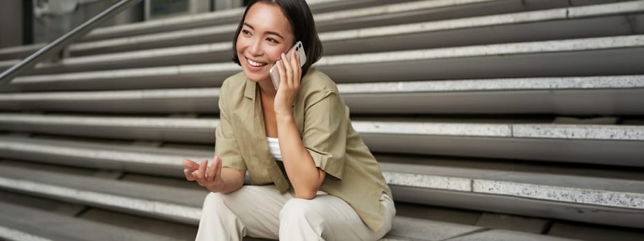 Close up portrait of smiling asian girl talks on mobile phone, sits outside on street stairs. Young woman calling friend on smartphone.